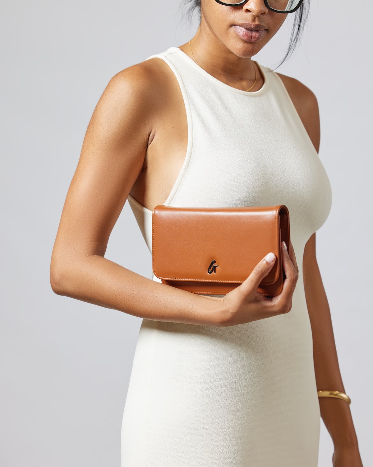 Woman holding a brown gunmetal WOC clutch bag with a black logo, dressed in a white sleeveless dress.
