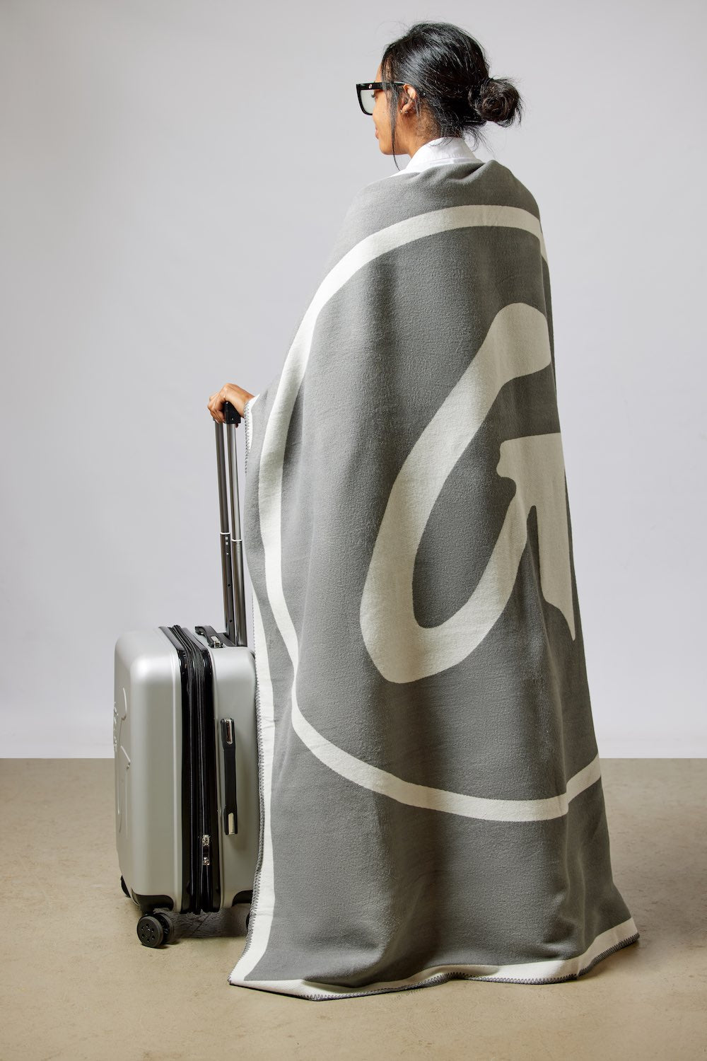 A woman with glasses and a bun, wrapped in a gray and white throw blanket, stands next to a silver suitcase in a minimal setting.