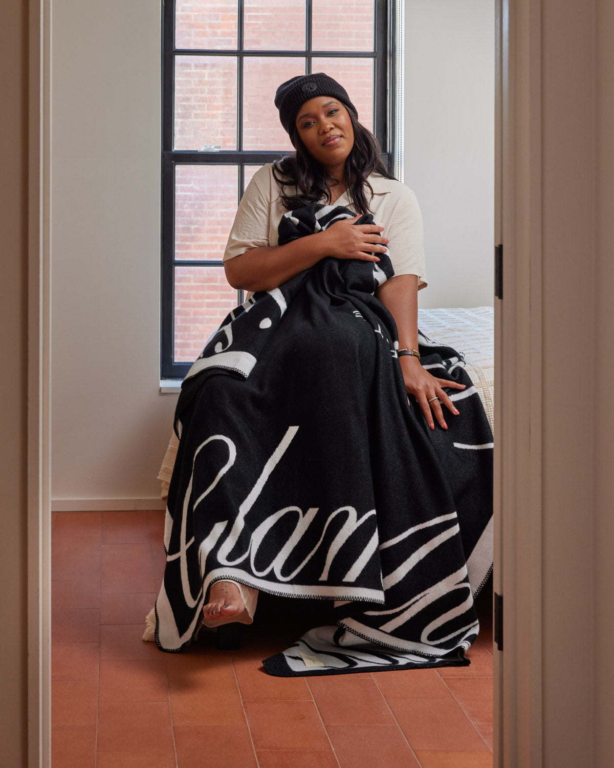 Woman sitting on bed wrapped in a black and white Signature Logo Luxe Throw Blanket, near a window with brick wall background.