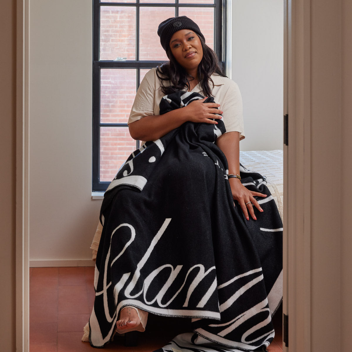 Woman sitting on bed wrapped in a black and white Signature Logo Luxe Throw Blanket, near a window with brick wall background.