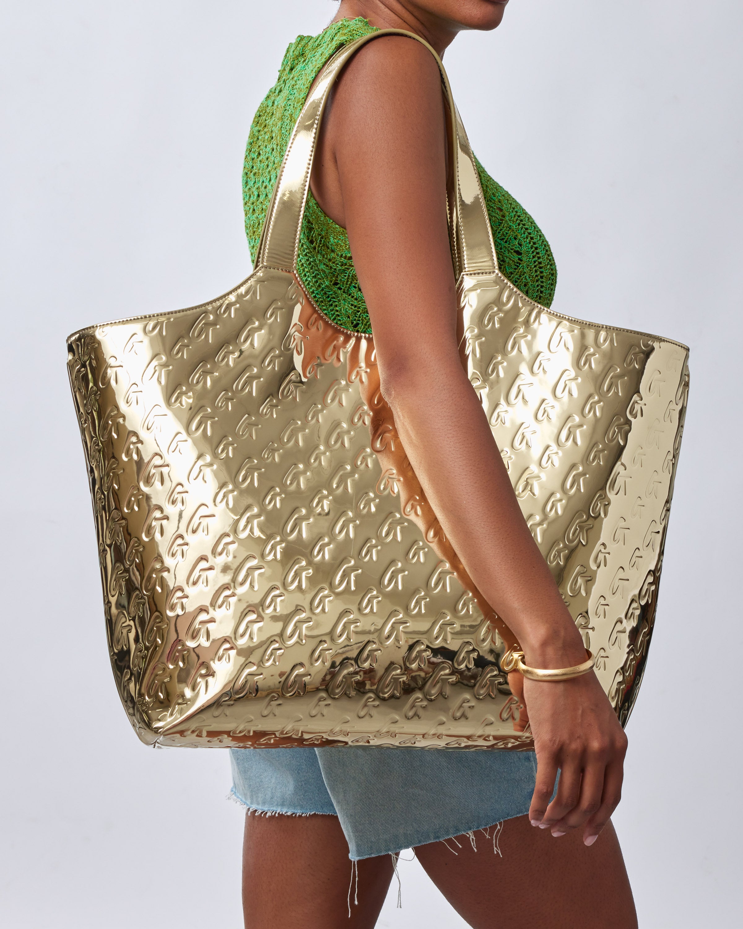 Woman holding a large metallic gold tote bag with embossed monogram pattern, paired with a green crochet top and denim shorts.