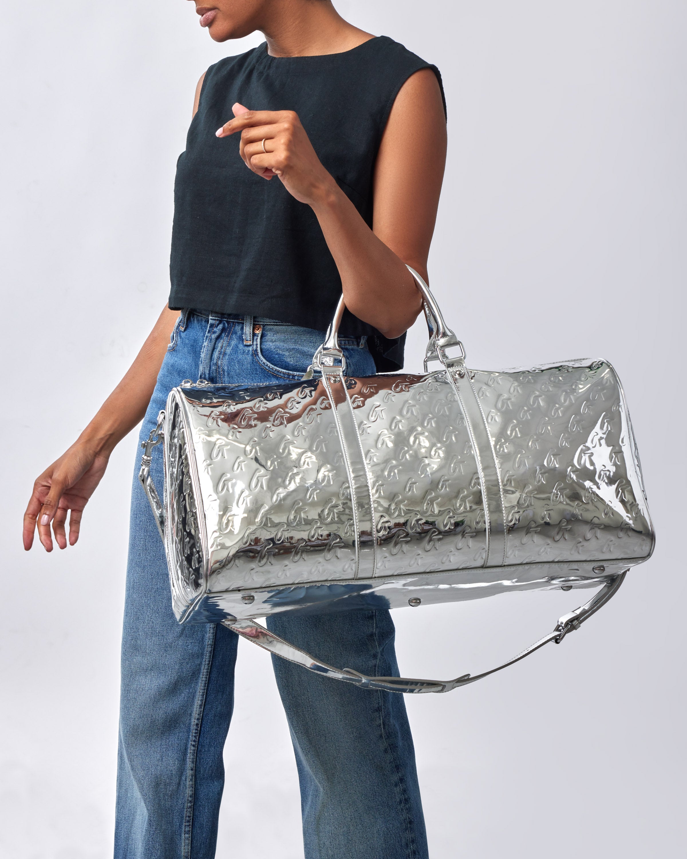 A woman holding a metallic silver duffle bag with embossed monogram pattern, silver handles, and adjustable strap.
