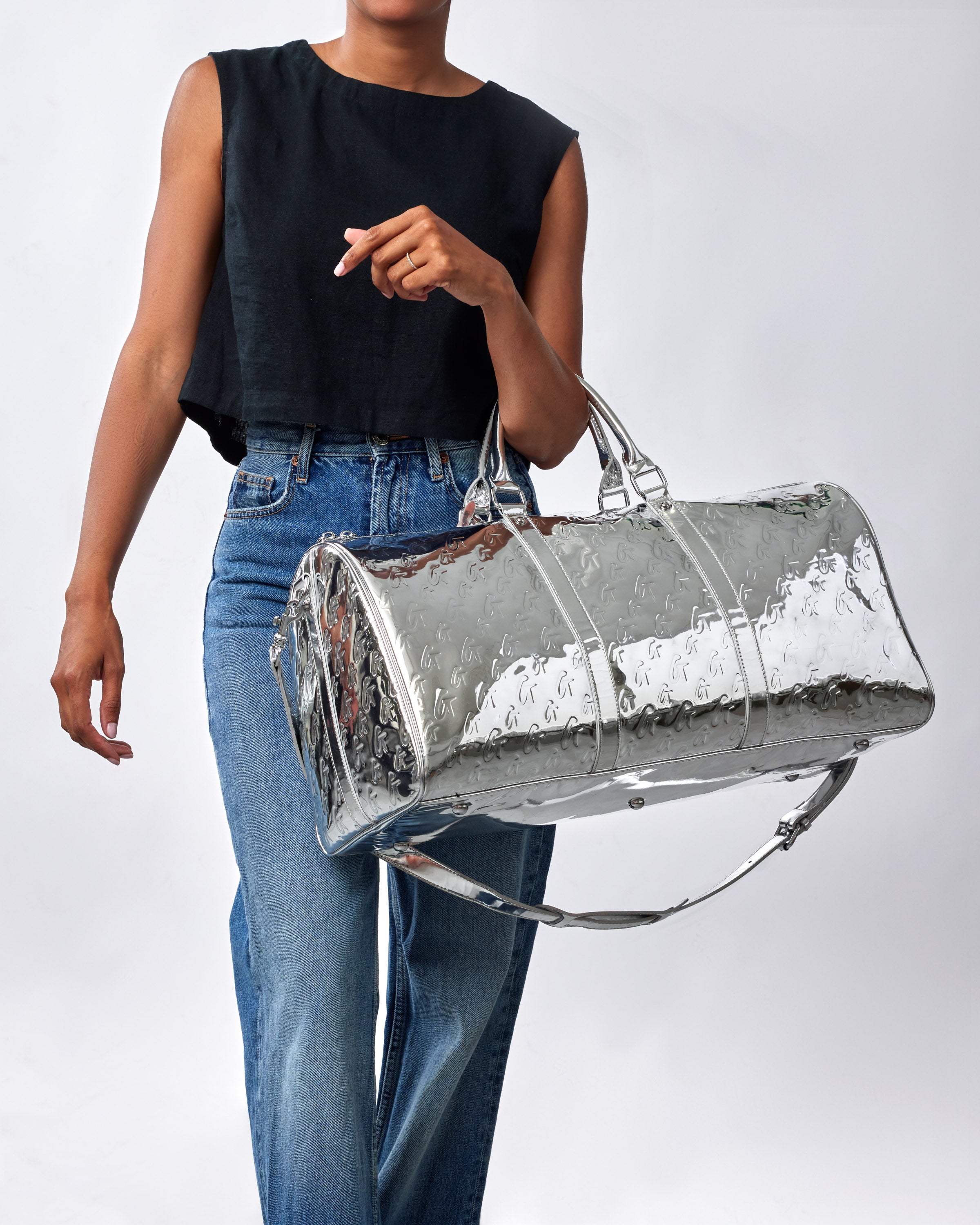 Metallic silver duffle bag with monogram pattern, shiny surface, and sturdy handles, carried by a woman in casual attire.