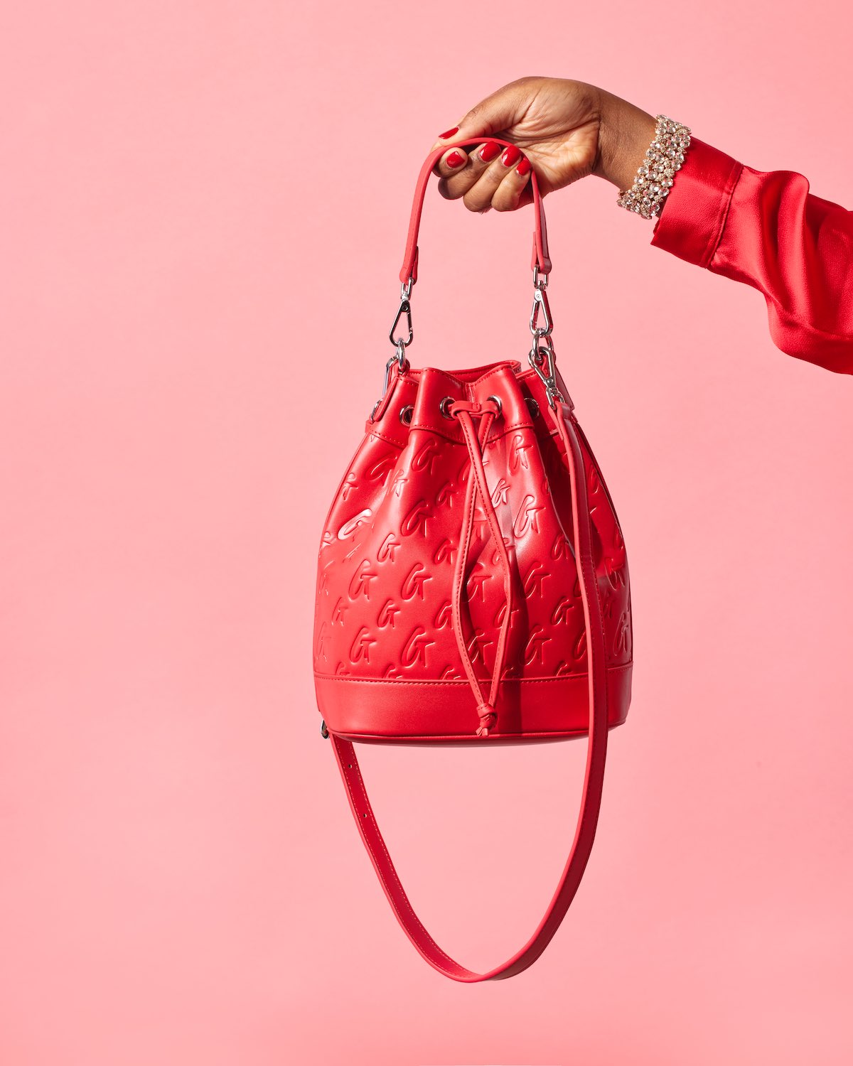 Red medium monogram bucket bag with embossed logo pattern, held by a hand with red nails and a pearl bracelet, against a pink background.