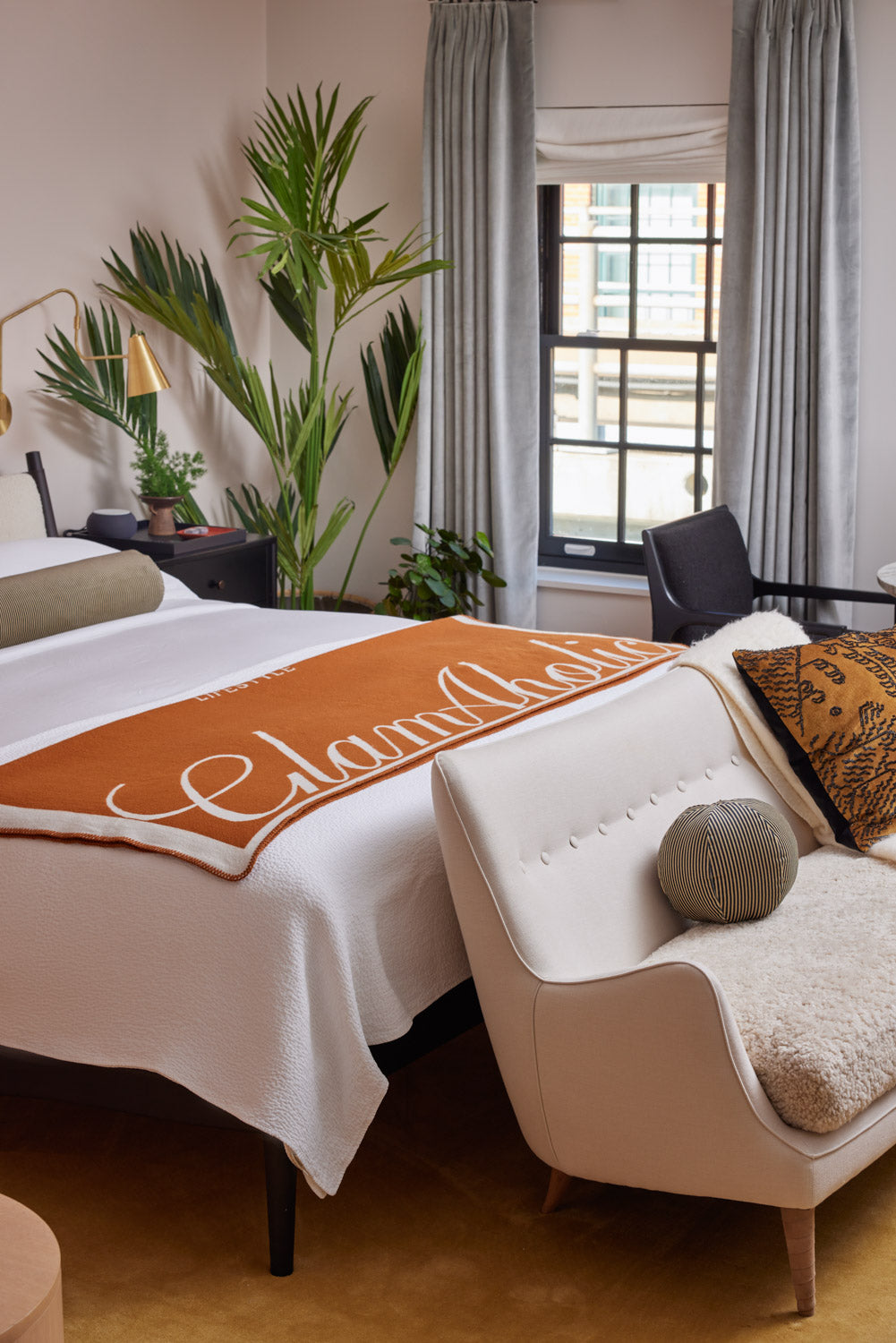Brown luxury throw blanket with white logo draped over a white bed in a stylish, cozy bedroom setting.