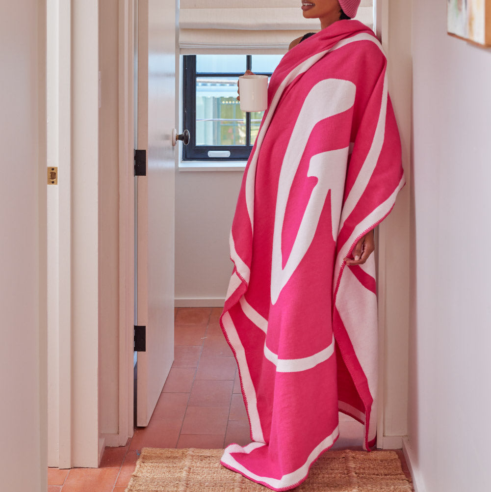 Woman wrapped in a bright pink and white throw blanket, wearing a matching pink beanie, standing in a cozy hallway.