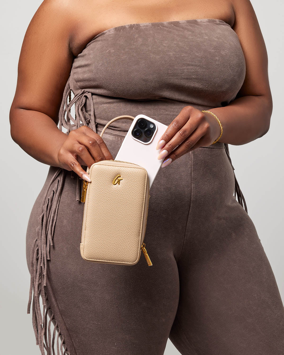Close-up of a woman holding a nude Pebble Phone Crossbody bag with a phone inside, against a neutral background.