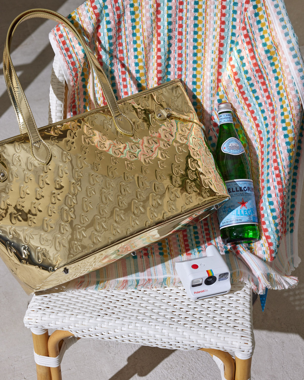 A shiny gold metallic tote bag with embossed monogram pattern, placed on a colorful striped towel on a white wicker chair.