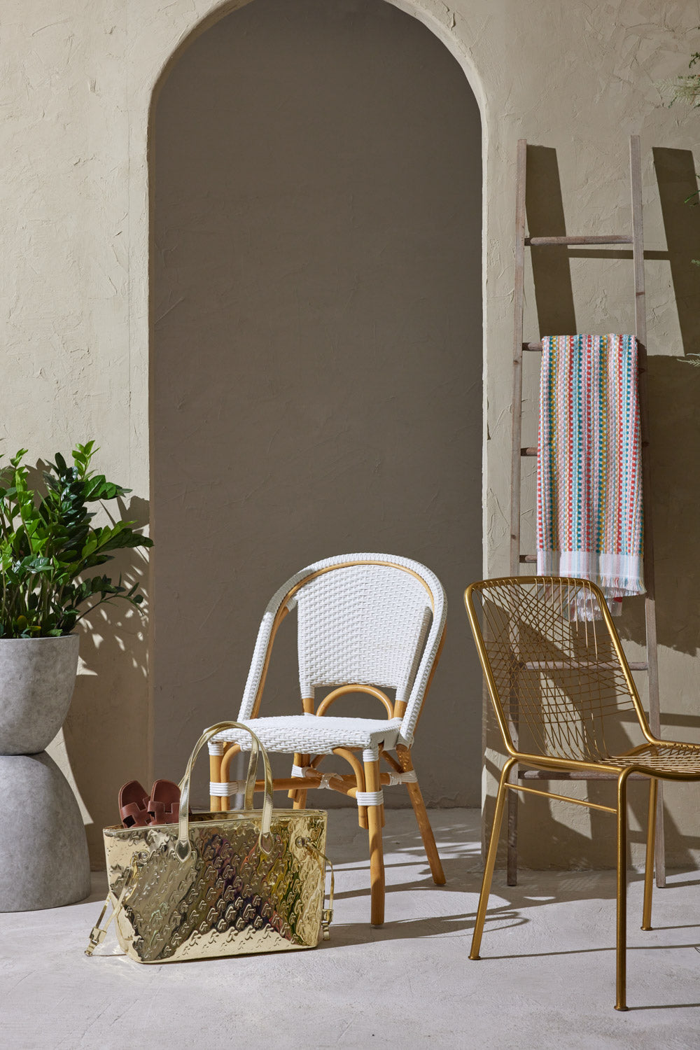 Metallic gold tote bag with monogram design, placed on the floor next to white and gold chairs in a stylish, modern setting.