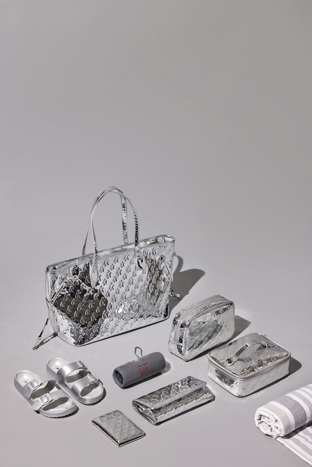 Set of metallic silver accessories including a tote, small pouch, wallet, sandals, JBL speaker, and striped towel.