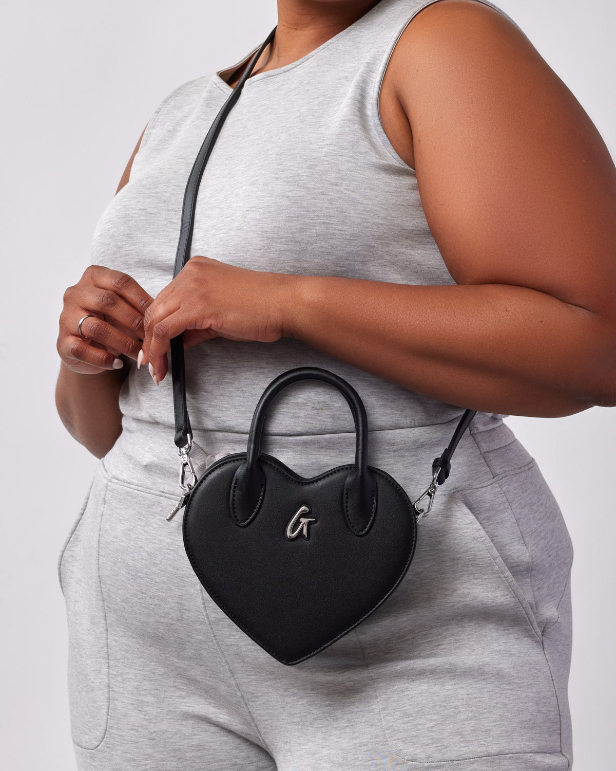 Black heart-shaped bag with a top handle, silver logo, and detachable shoulder strap, worn by a person in gray athletic wear.