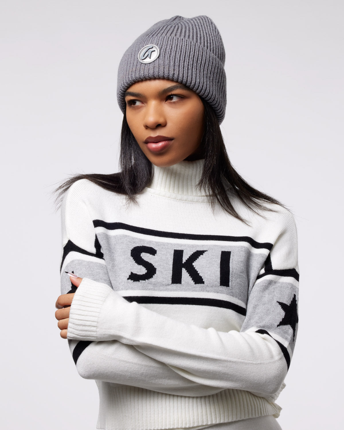 Gray knit beanie with a logo patch, worn by a woman in a white sweater with "SKI" and star designs.