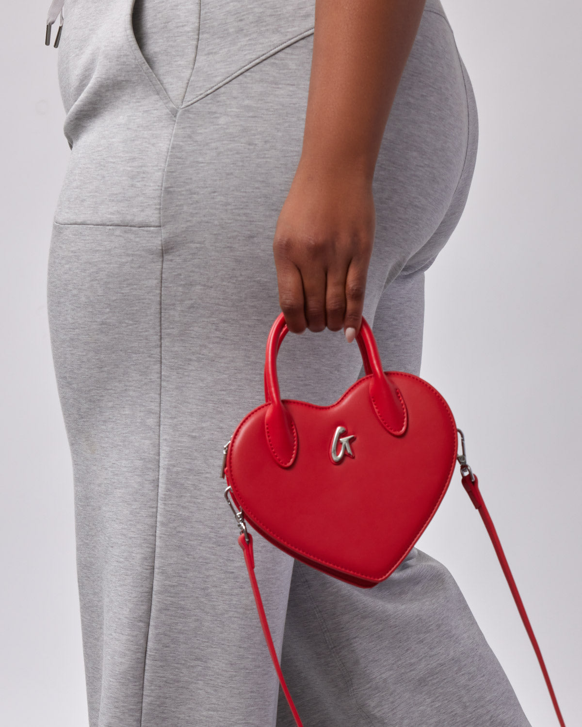Red heart-shaped handbag with silver logo, top handle, and adjustable shoulder strap, against gray casual wear.