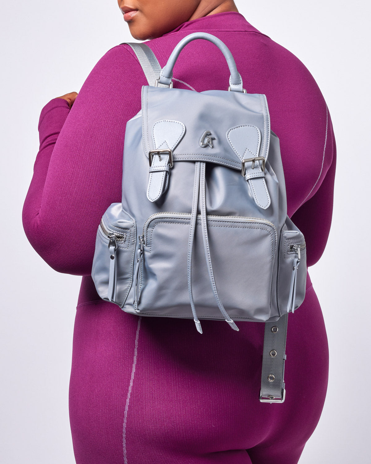 Light gray designer-style backpack with front zipper pockets, buckle details, and silver hardware, worn by a woman in a ma...