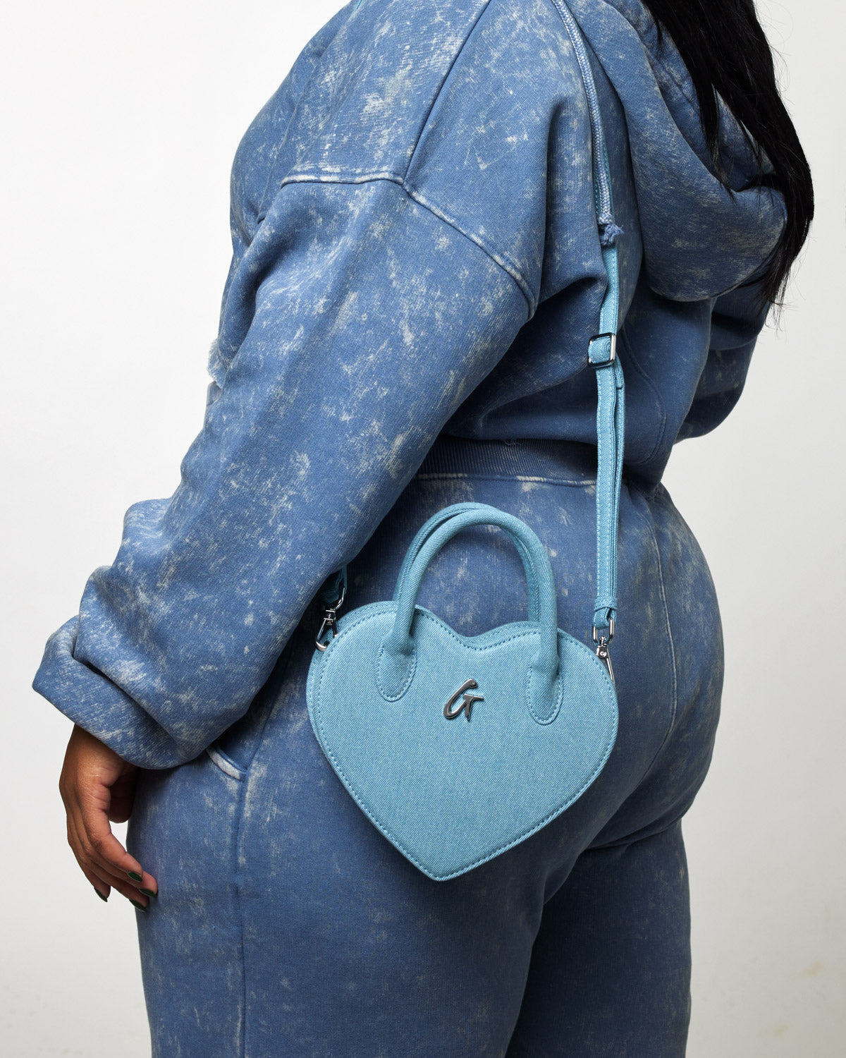 A person wearing a matching denim hoodie and jeans carries a light blue heart-shaped denim bag with a small handle and a strap.