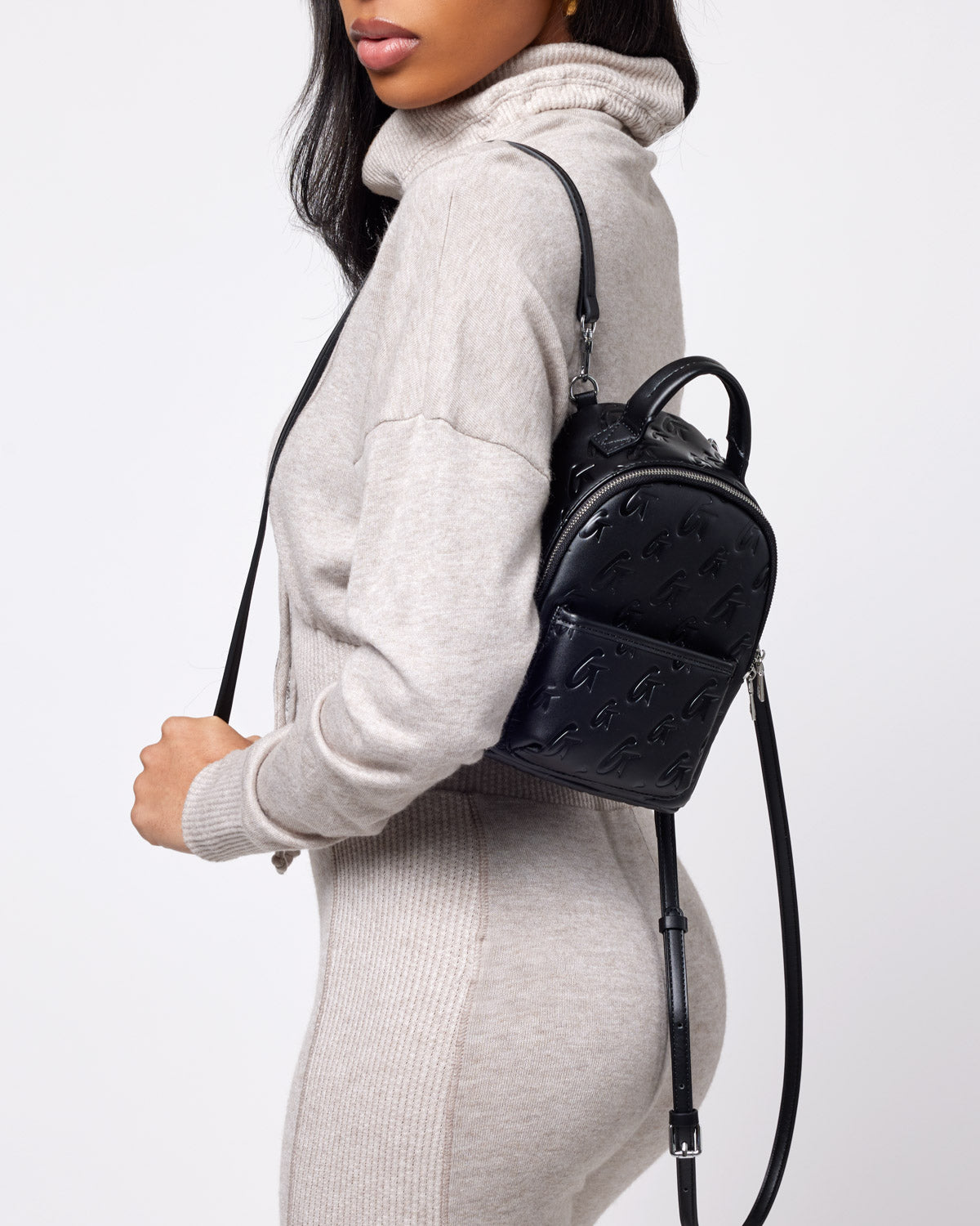 Black mini monogram backpack with embossed logo pattern, top handle, adjustable strap, and front zip pocket, worn by a woman.