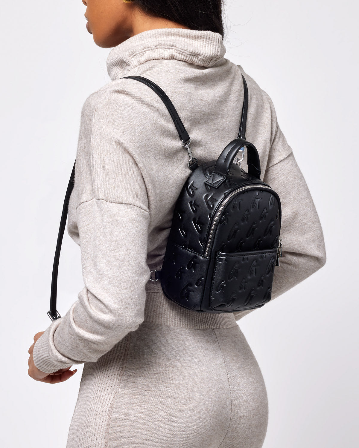 Small black Monogram backpack with embossed logo pattern, carried by a woman in a beige hoodie and matching pants.