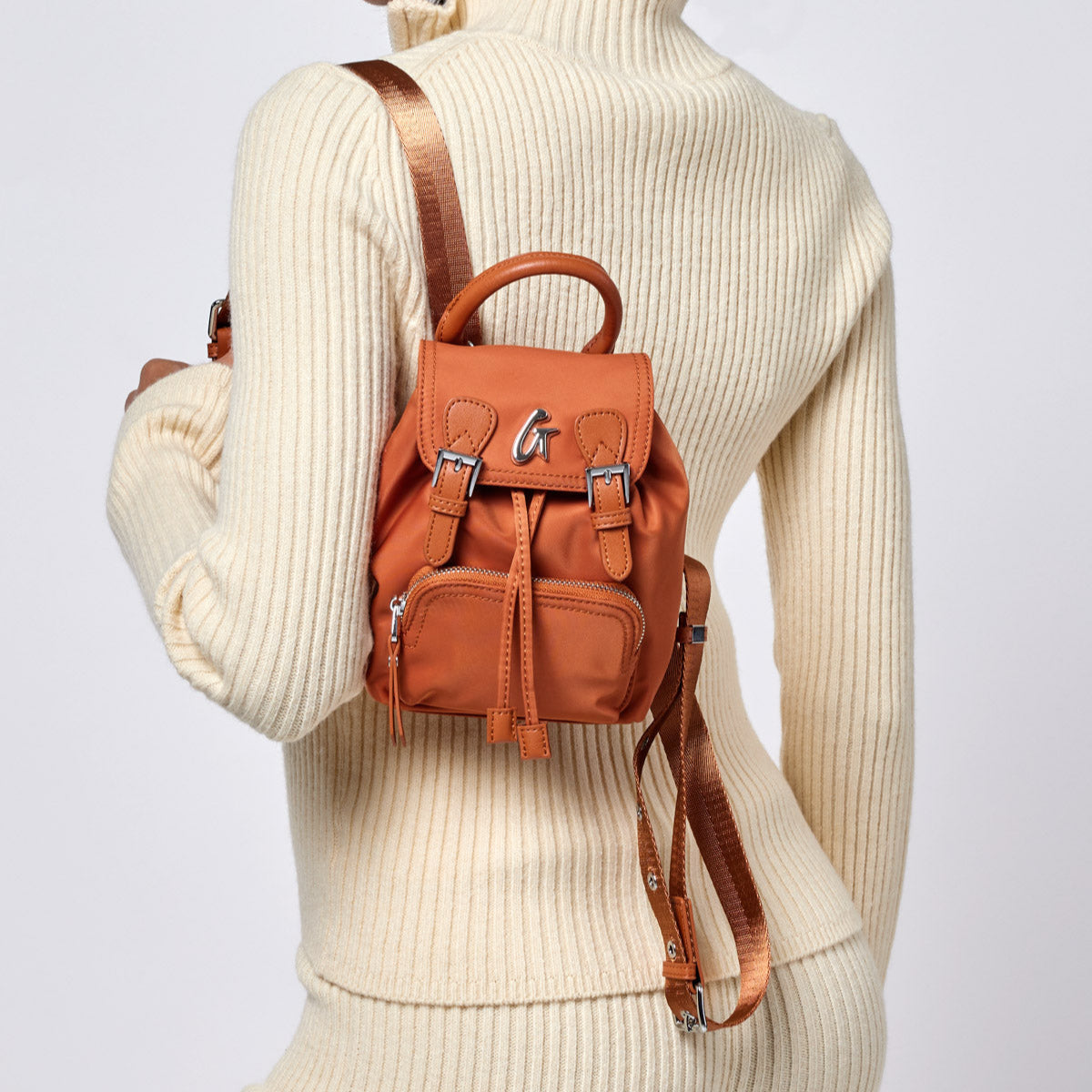 Small brown nylon backpack with a top handle, front zipper pocket, adjustable straps, and metal logo detail.