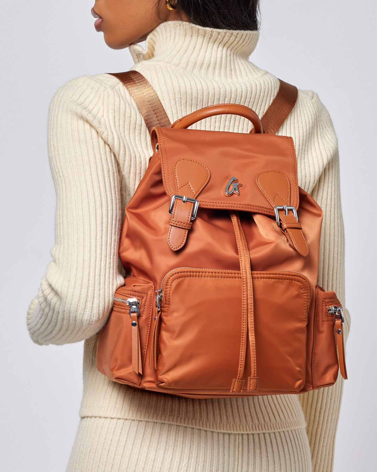 Brown nylon backpack with multiple zippered compartments, adjustable straps, and a top handle, worn over a cream turtleneck sweater.