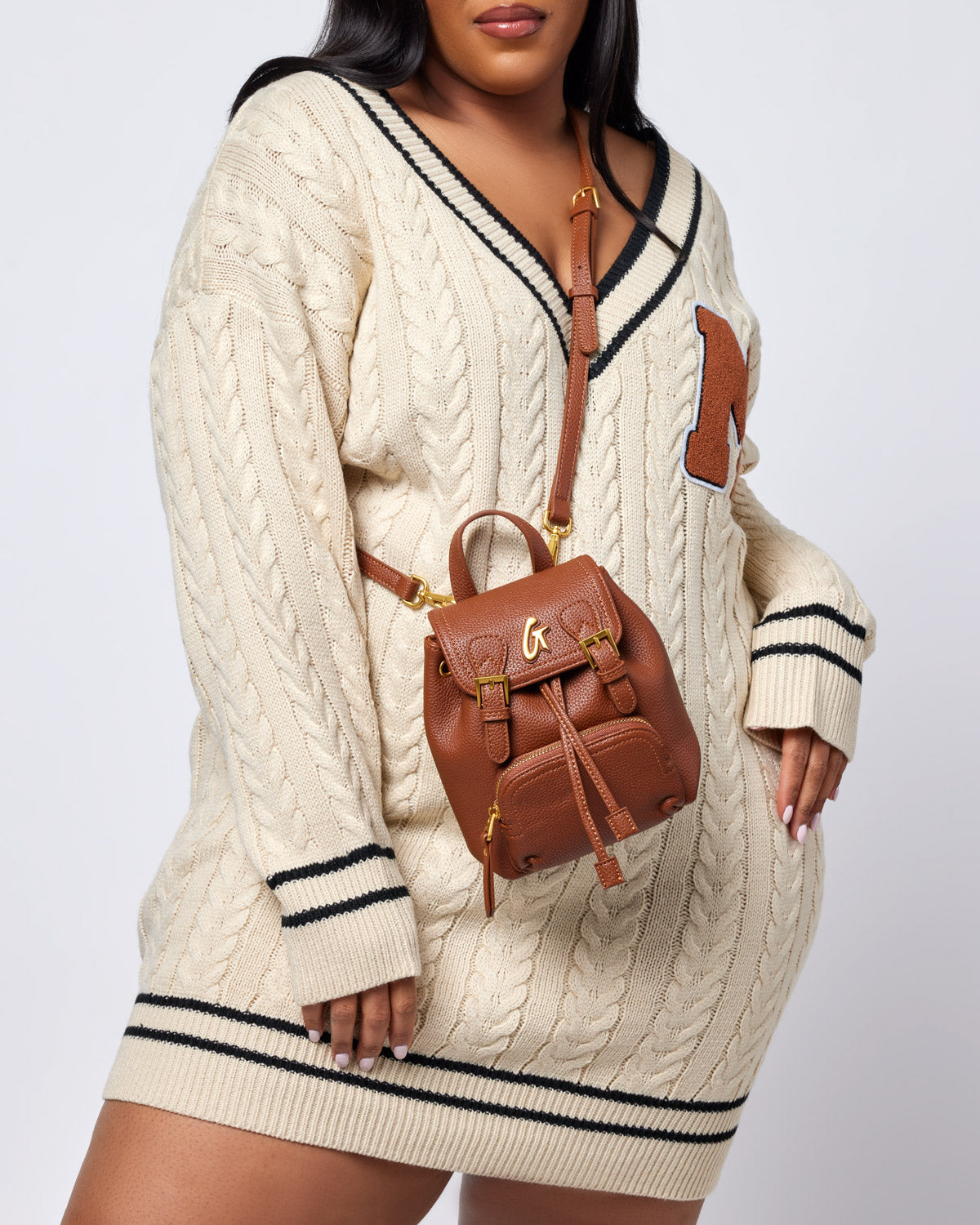Brown mini pebble crossbody backpack with gold hardware, worn over a cream cable-knit sweater.