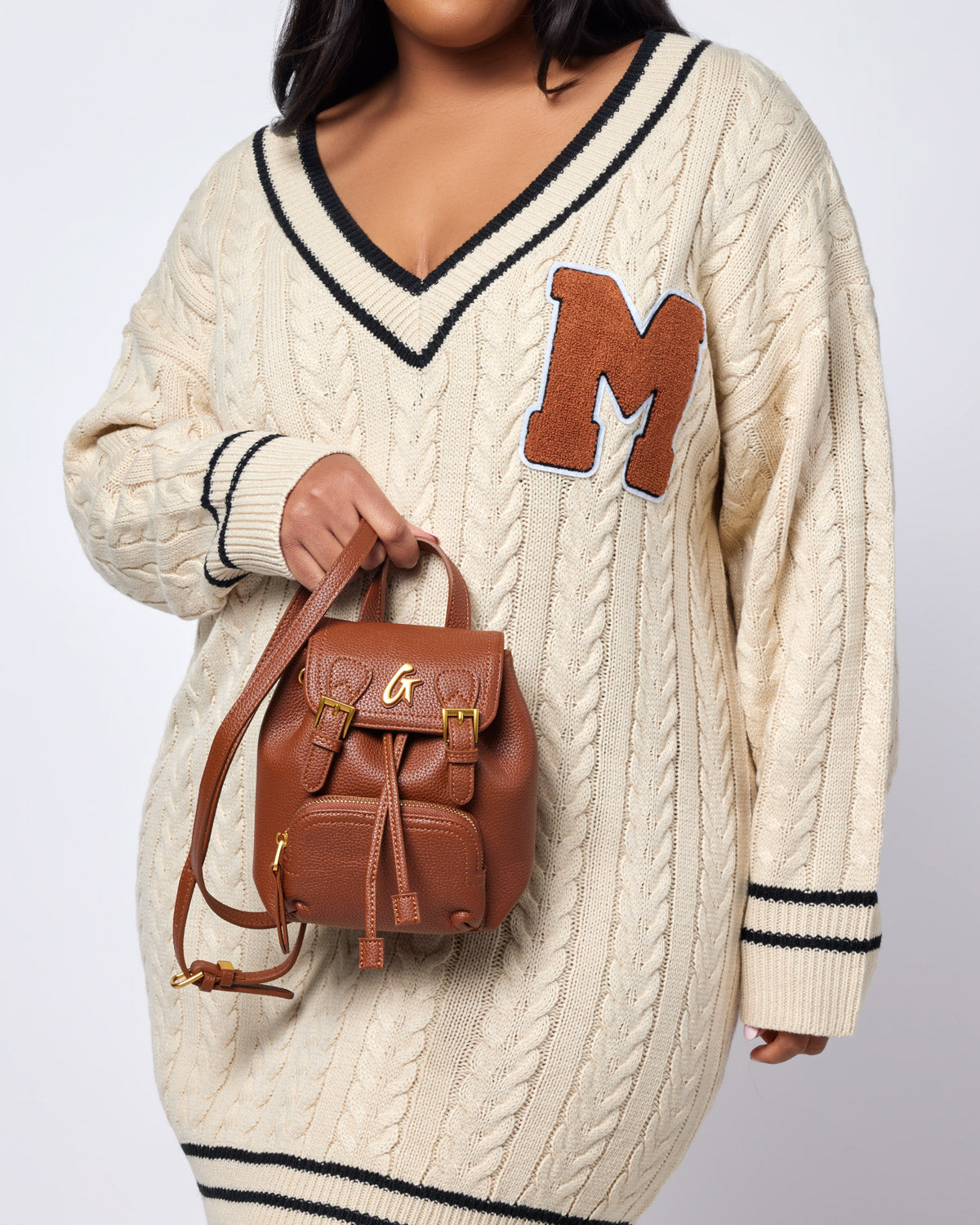 Brown mini pebble crossbody backpack with gold hardware, worn over a cream cable-knit sweater with black accents.