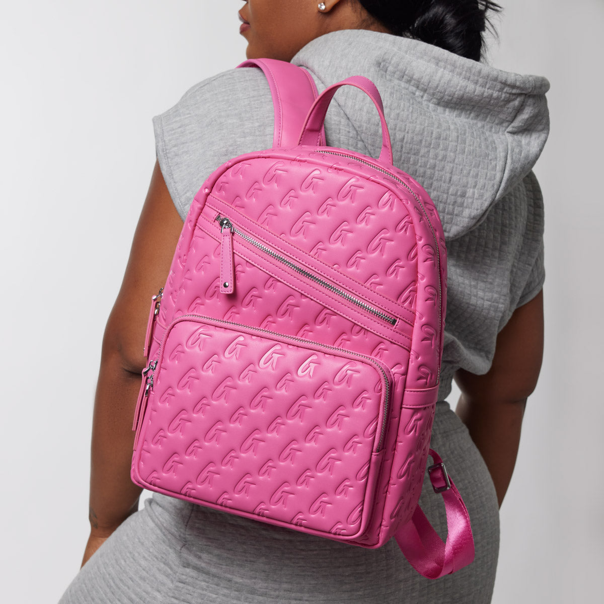 Back view of a woman wearing a grey hoodie and a matching grey skirt, carrying a vibrant hot pink monogram backpack with embossed pattern.