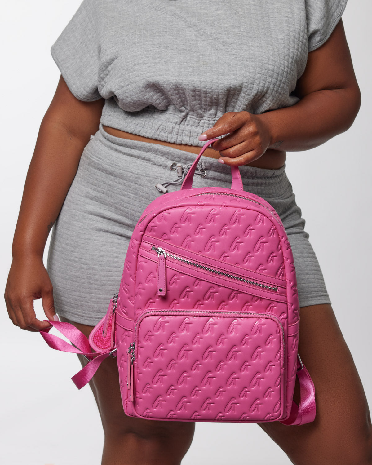 A woman holds a bright pink Monogram Backpack with embossed logo pattern, multiple zippered compartments, and adjustable straps.