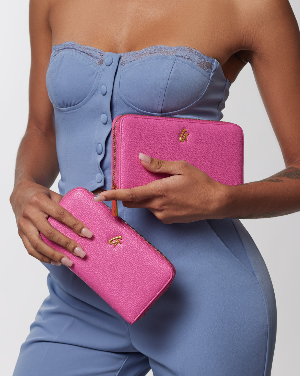 A woman in a blue outfit holds a hot pink pebble-textured wallet, a matching zippered pouch, and a larger pink clutch with gold accents.