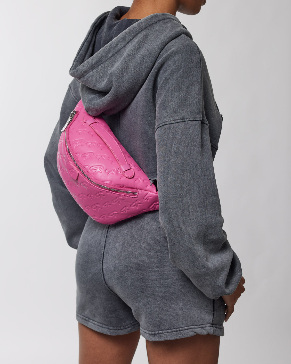 Pink monogram Fanny Pack with embossed logo, worn crossbody over a gray hoodie and shorts.