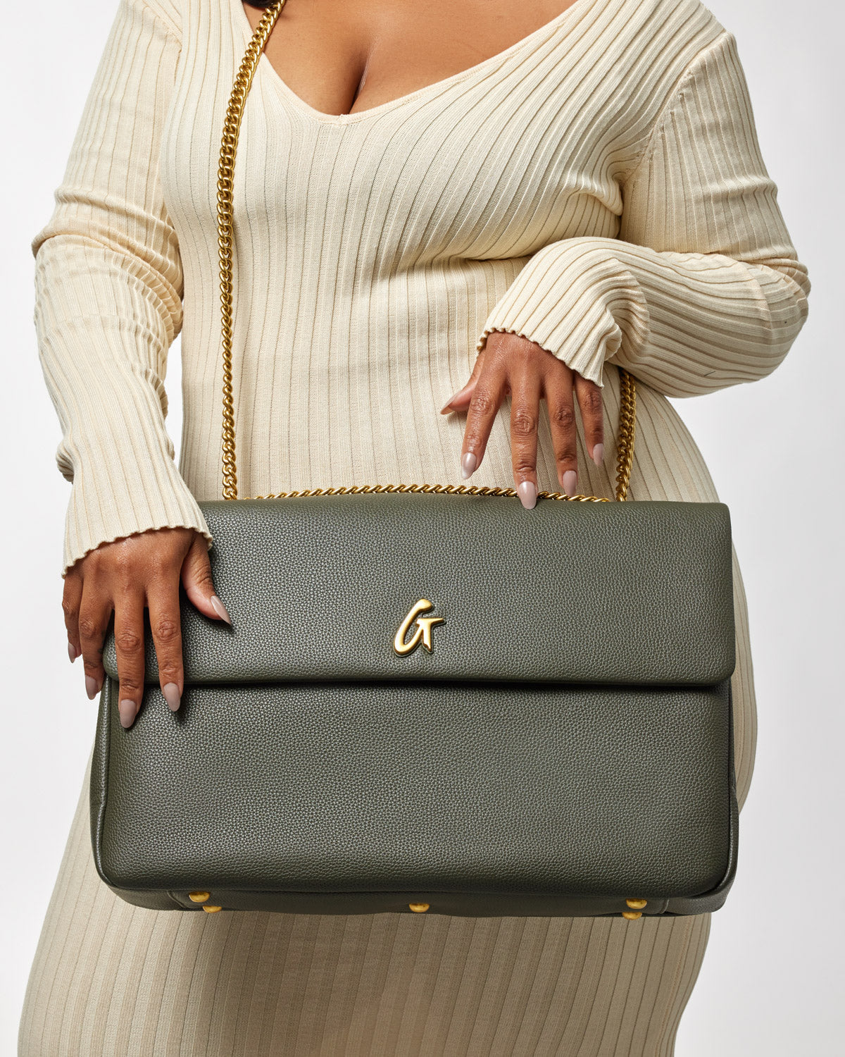 Olive green XL Pebble Flap Bag with gold chain strap and logo, held by a woman in a cream ribbed outfit.