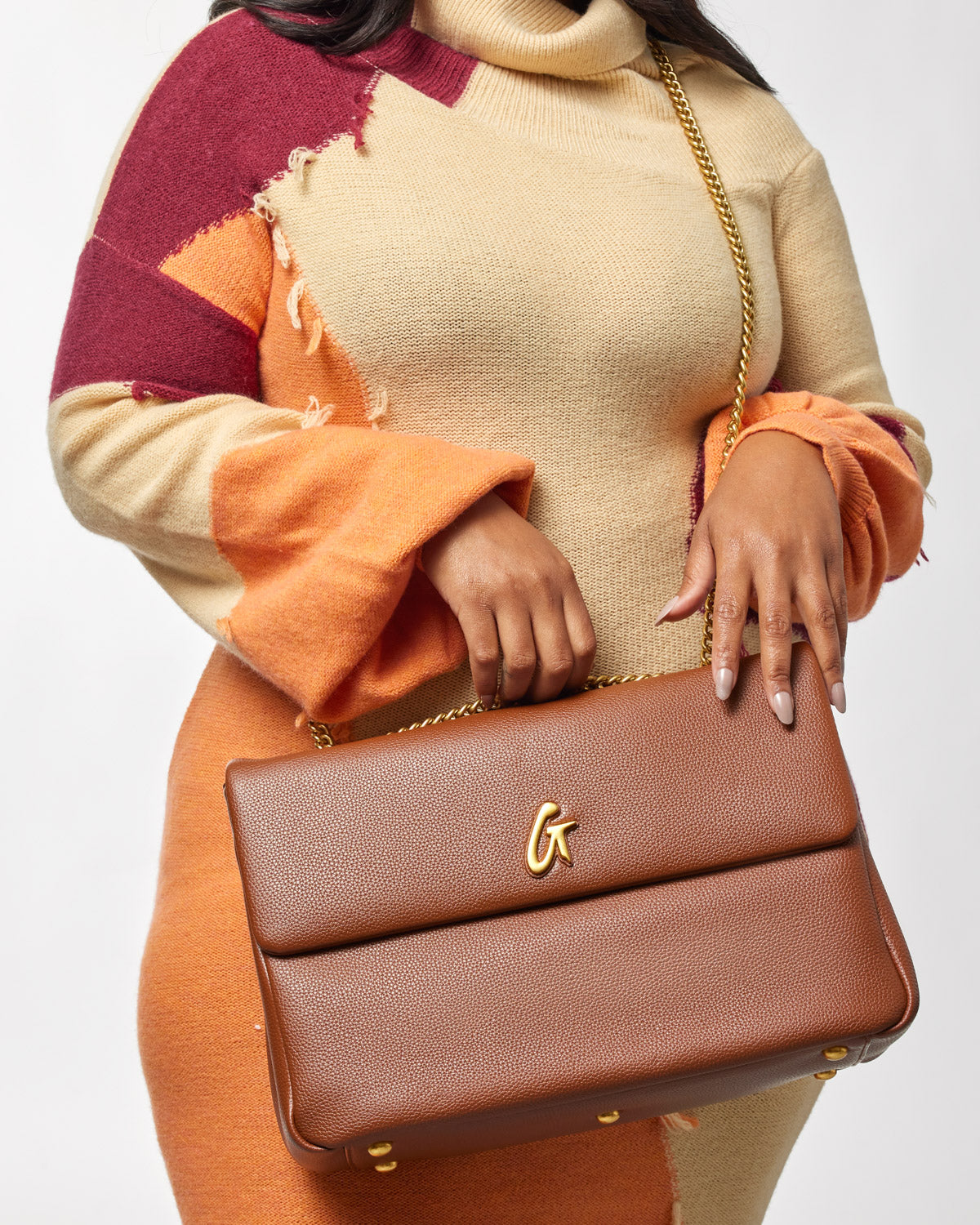 Large pebble flap bag in brown with a gold chain strap and a gold logo on the front, held by a woman in a colorful sweater.