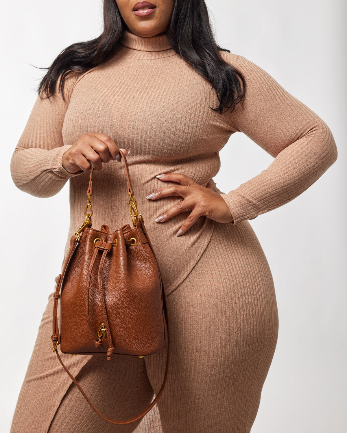 Medium brown pebble bucket bag with a drawstring closure, gold hardware, and a short handle, held by a woman in a beige outfit.