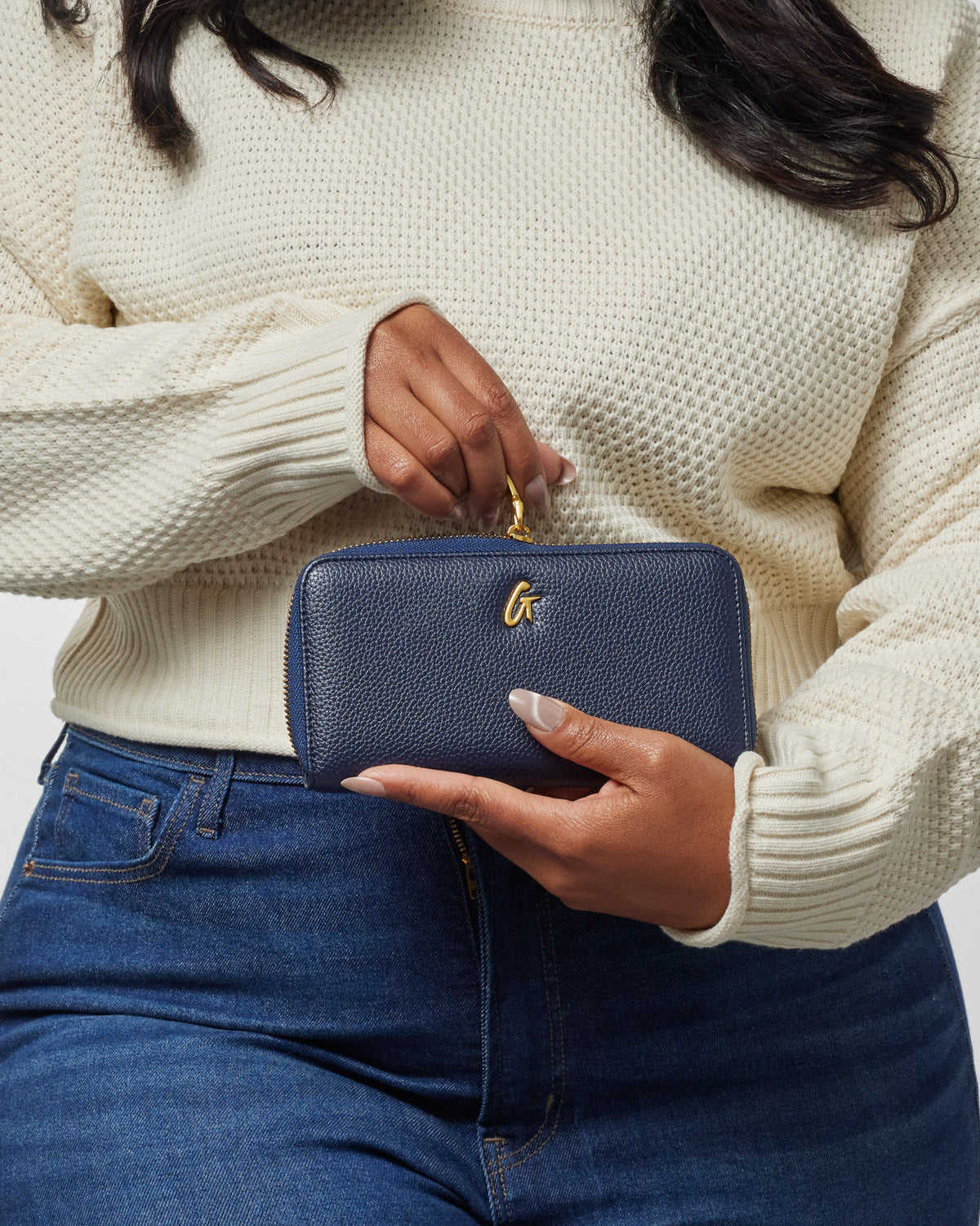 Person holding a navy pebble leather wallet with a gold "G" logo, gold zipper, and a top handle, against a casual outfit.