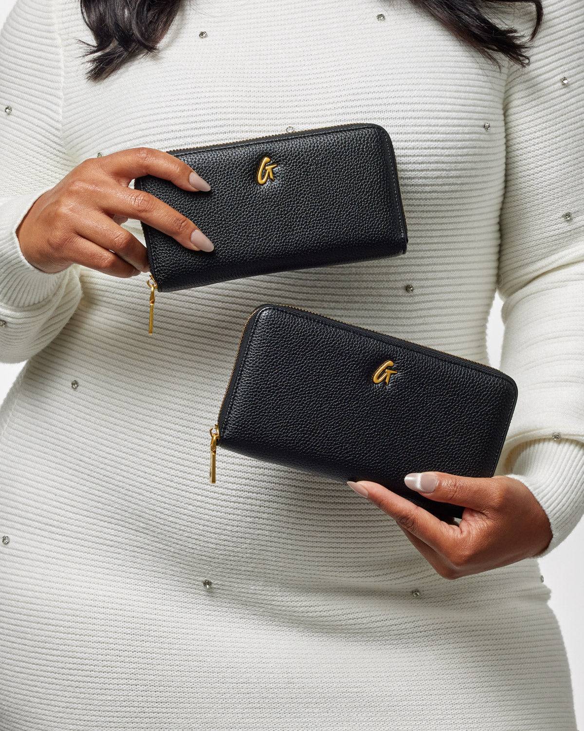 Two black textured wallets with gold zippers and a gold "G" logo, held by a person in a white, jeweled sweater.