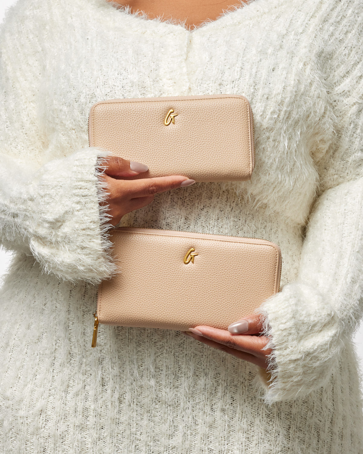 Two beige pebble-textured wallets with gold "G" logos, held by a person in a cozy white sweater.