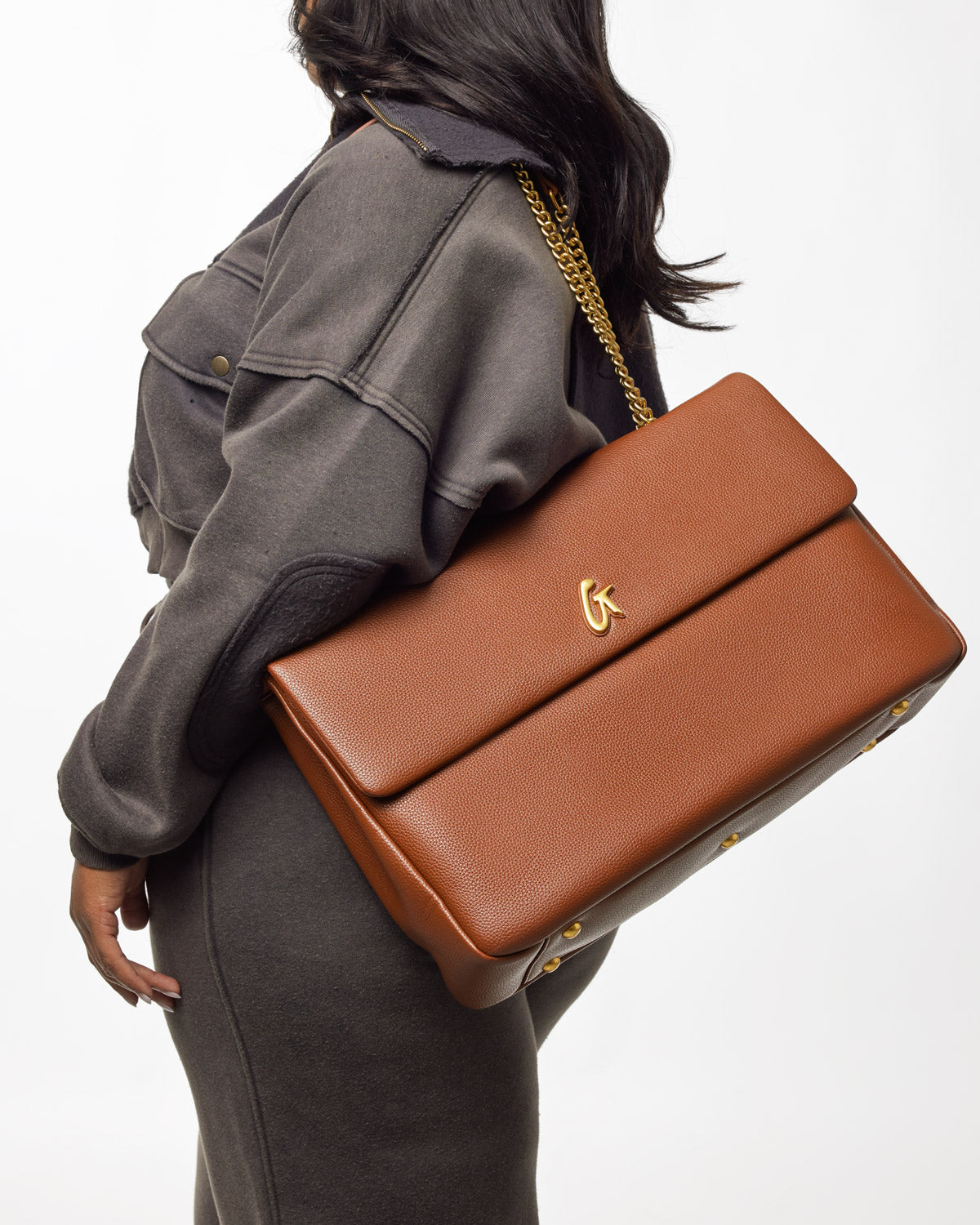 Large brown pebble leather flap bag with gold chain strap and logo detail, carried over a woman's shoulder.