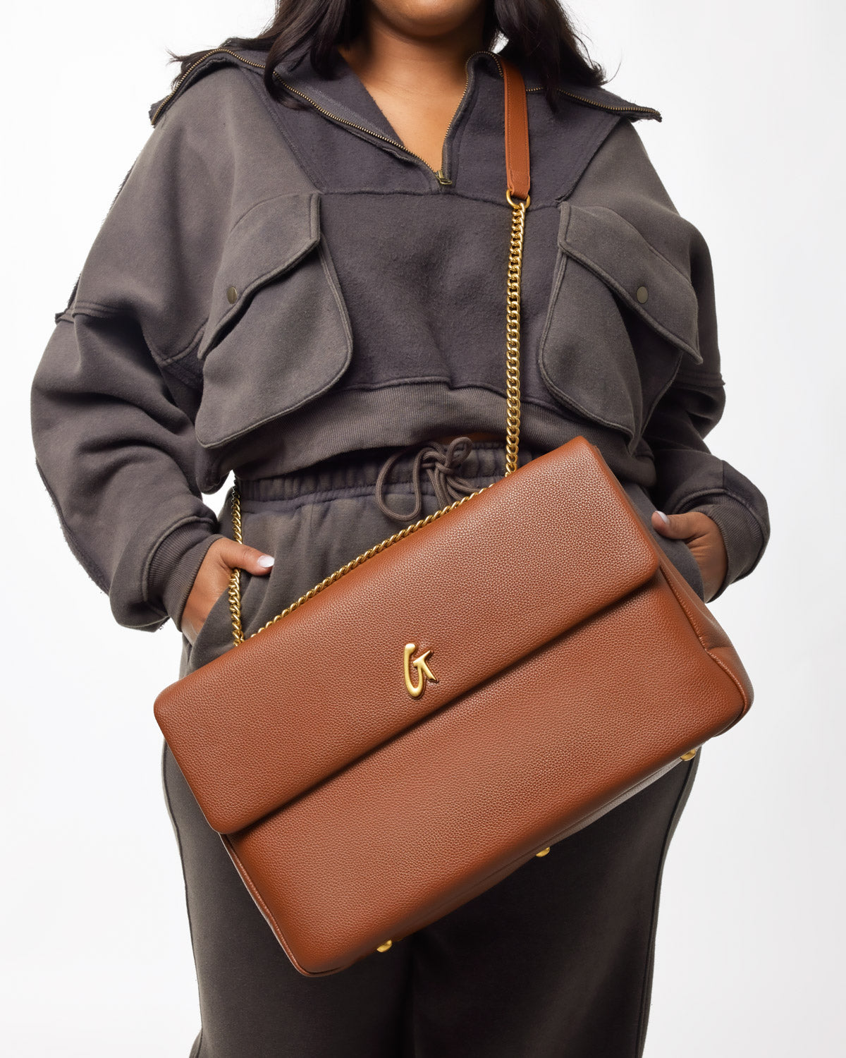 Large brown pebble-textured flap bag with gold chain strap and logo accent, held by a person wearing a gray cropped jacket.
