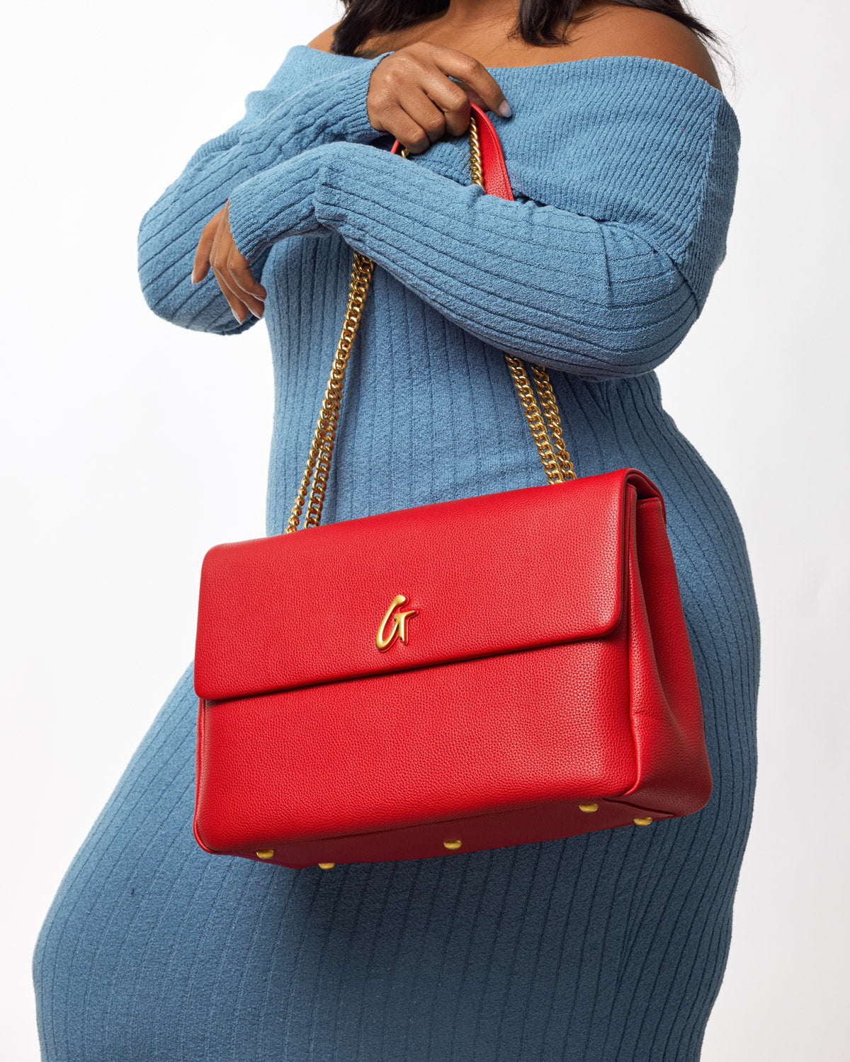 Red large pebble flap bag with gold chain strap and logo, carried by a woman in a blue ribbed sweater.