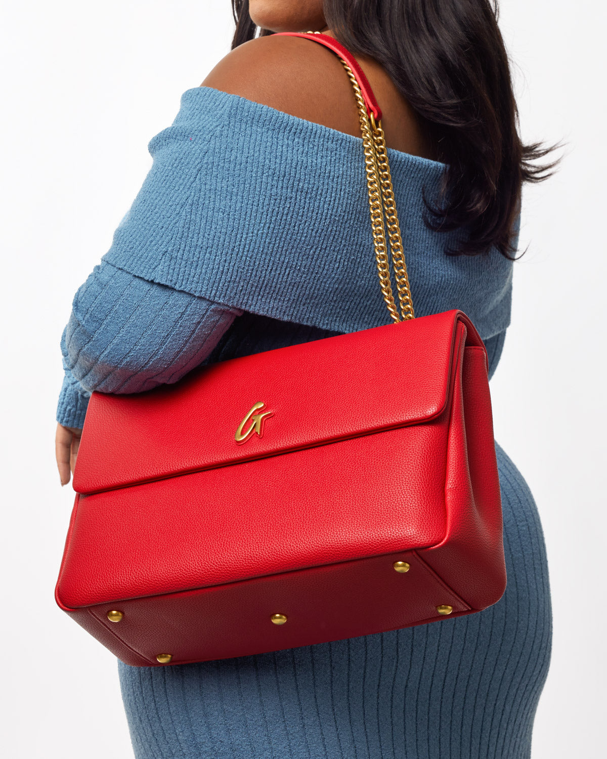 Large red pebble flap bag with gold chain strap and gold logo, worn over shoulder by a woman in a blue off-shoulder sweater.