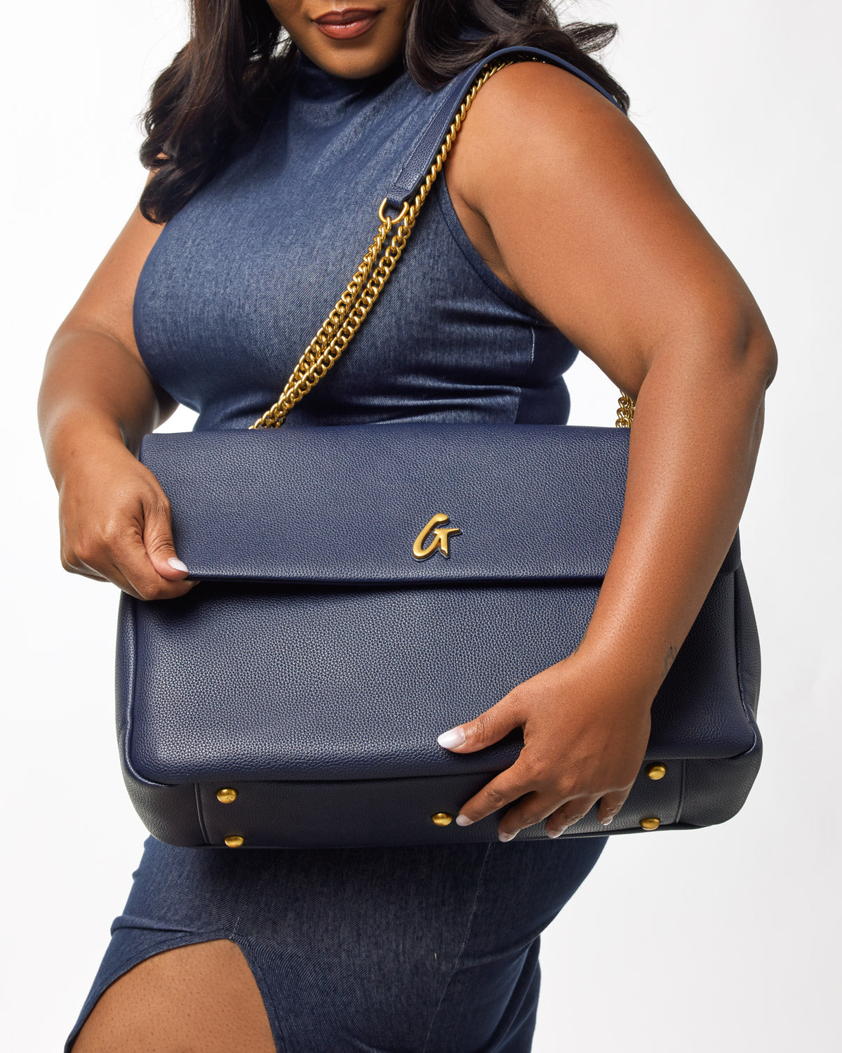 A woman holds a navy XL Pebble Flap Bag with gold chain strap and gold logo, featuring textured leather and gold accents.