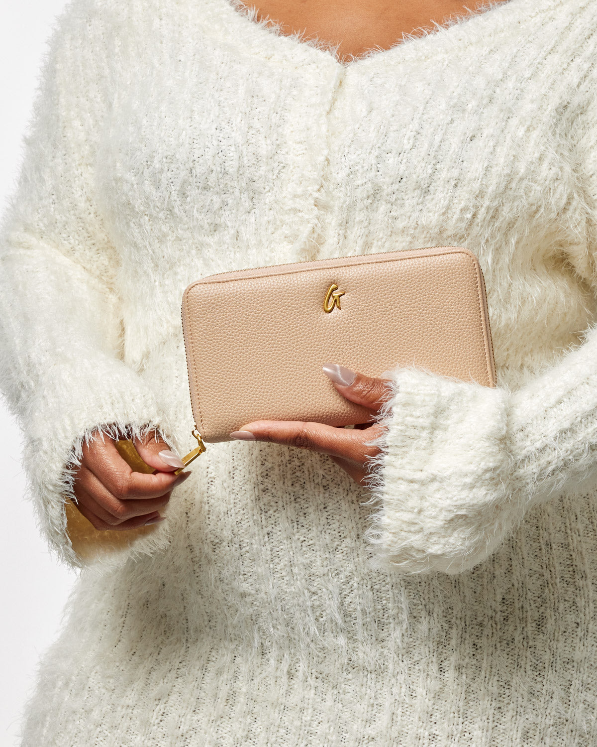 A person wearing a white fuzzy sweater holds a nude-colored jumbo pebble wallet with a gold logo and zipper.