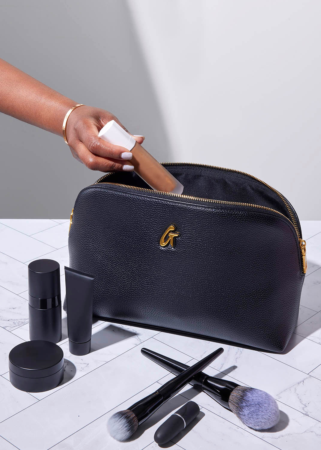 Black jumbo pebble cosmetic pouch with gold zipper, holding makeup products and brushes on a marble surface.