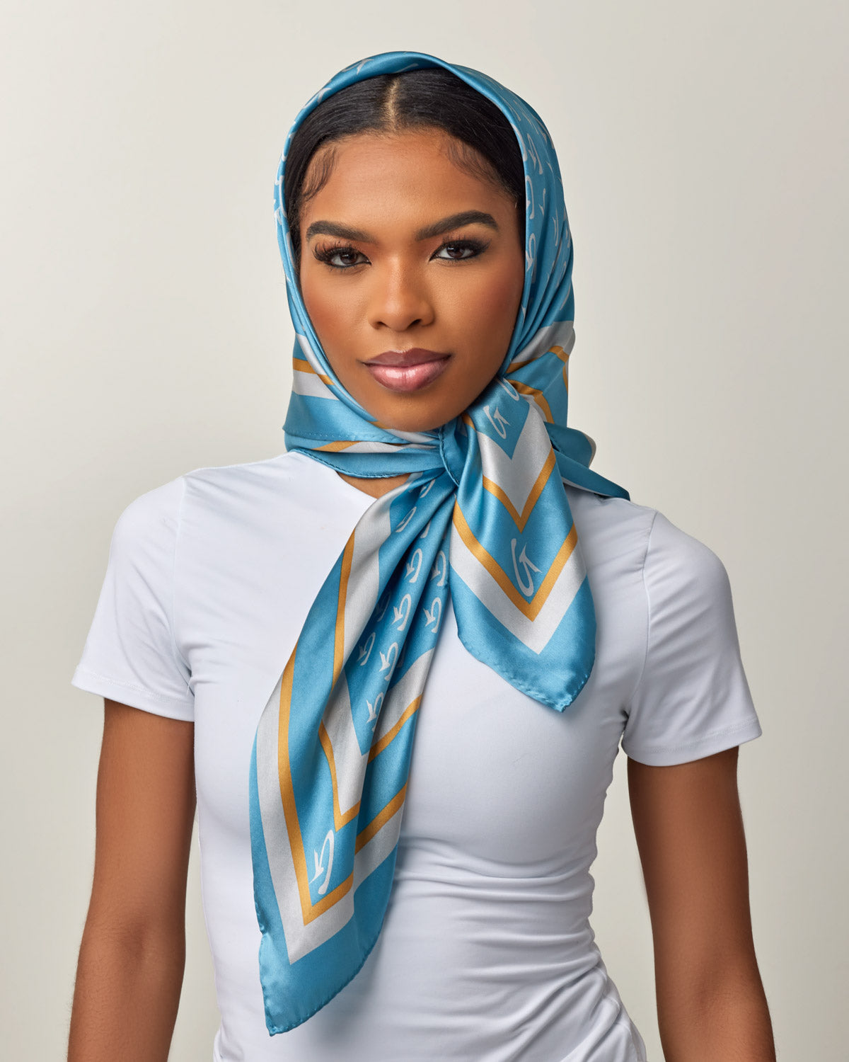 A woman wears a large, blue silk scarf with gold and white monogram patterns, styled as a headscarf over a white top.