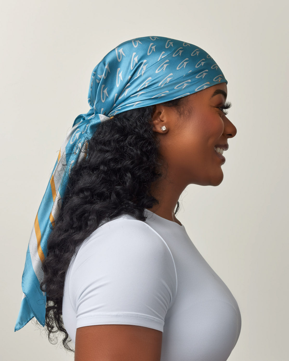 A woman wearing a large blue silk scarf with gold monogram pattern, tied at the back, paired with a white top.