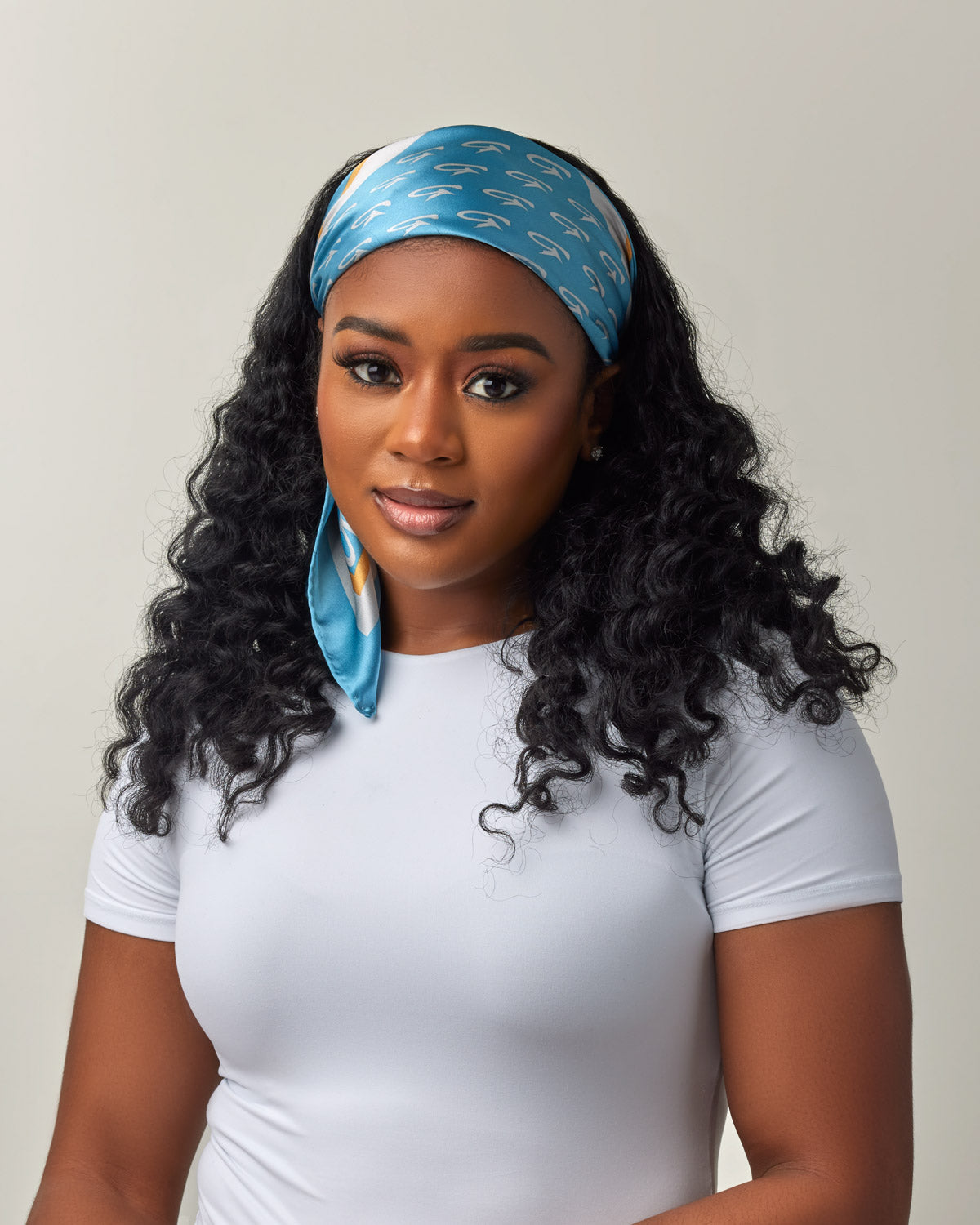Woman wearing a blue and gold silk scarf with monogram pattern, styled as a head wrap, against a neutral background.