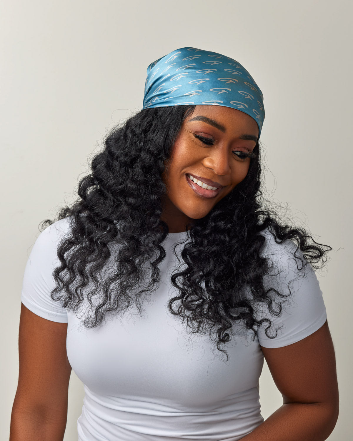 Woman wearing a blue silk scarf with monogram pattern, styled as a head wrap, paired with a white top.