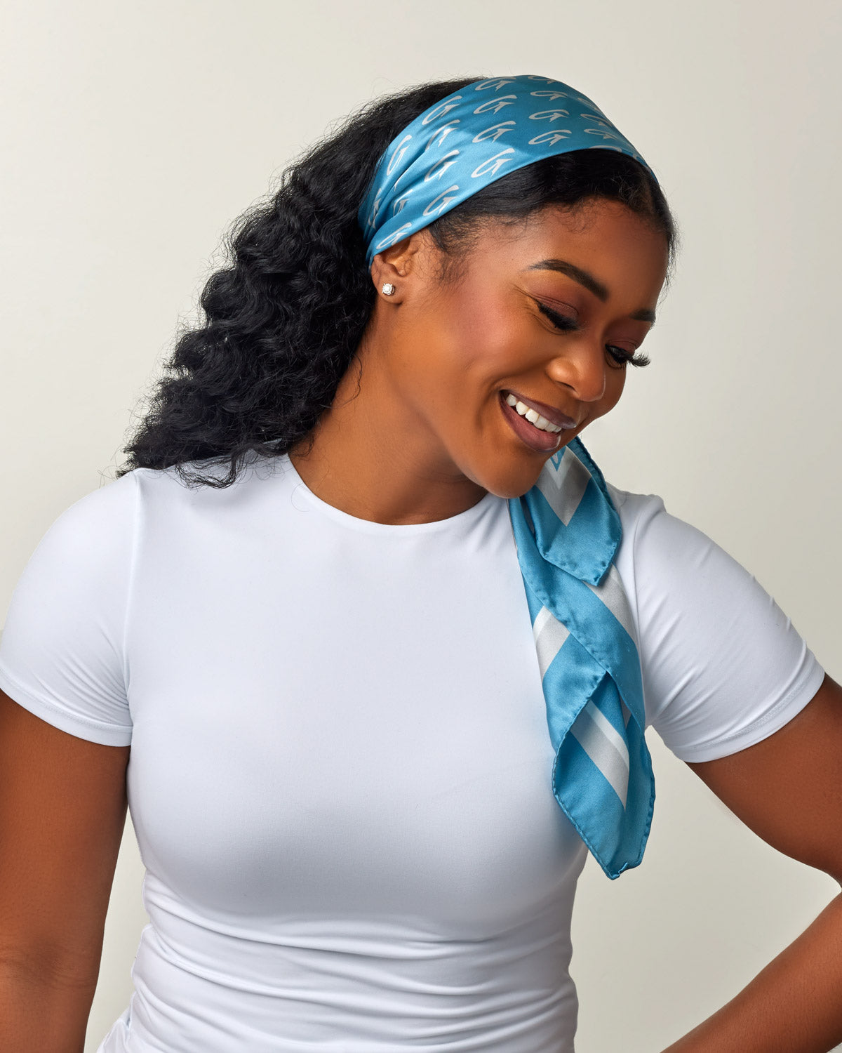 Woman wearing a blue silk scarf with silver monogram pattern, styled as a headband, over a white t-shirt.