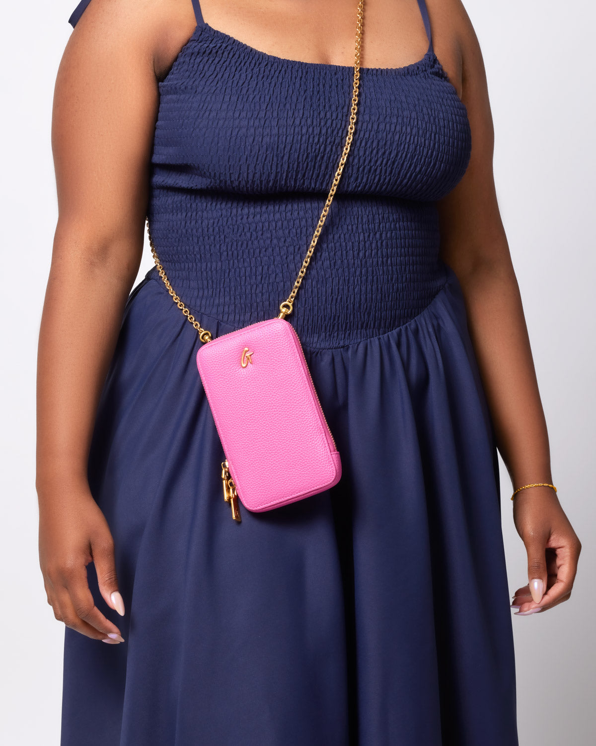 Pink Pebble Phone Crossbody bag with gold chain strap worn across a woman's shoulder, paired with a navy blue dress.
