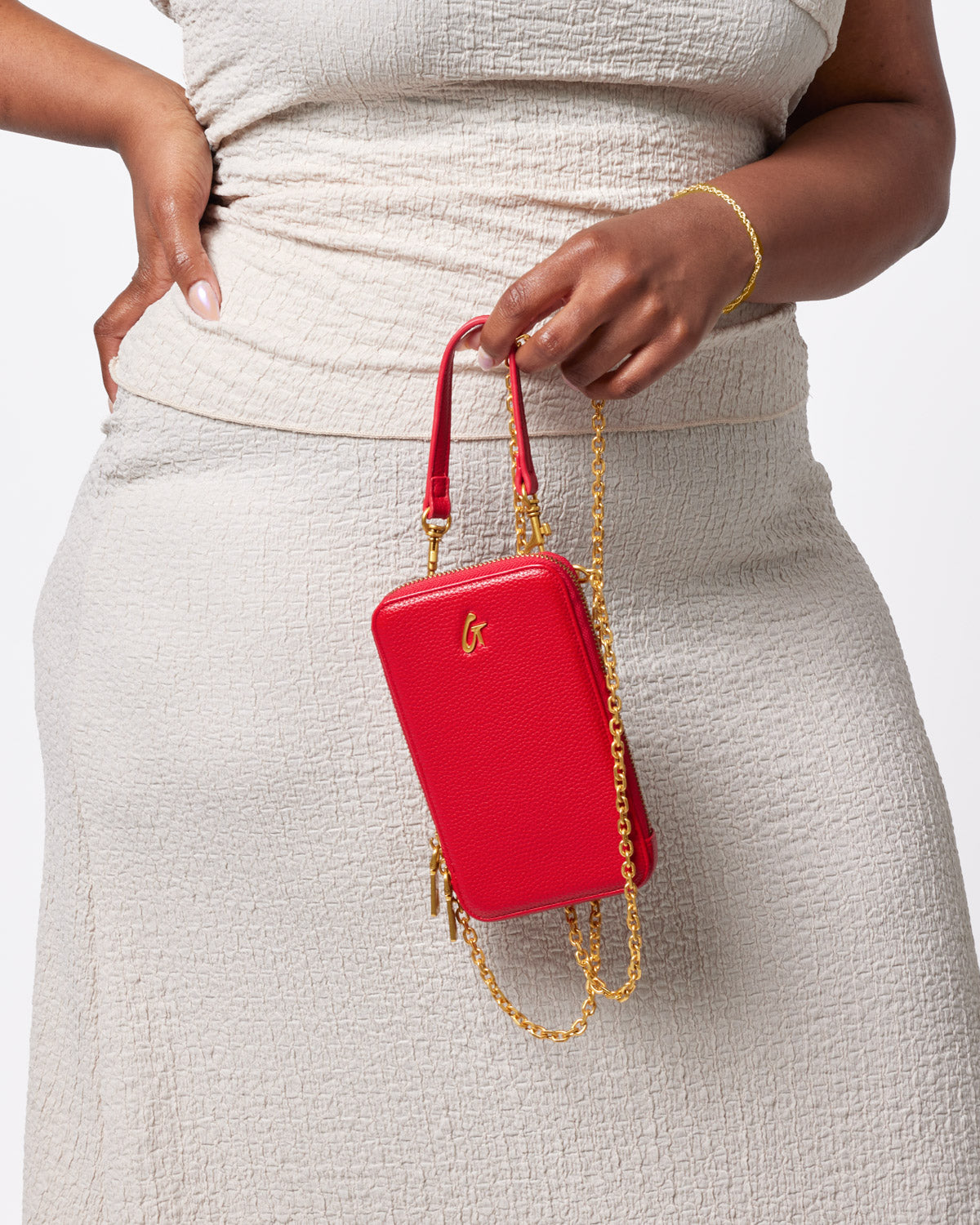 Red pebble-textured crossbody phone bag with gold chain strap and logo detail, held by a person in beige clothing.