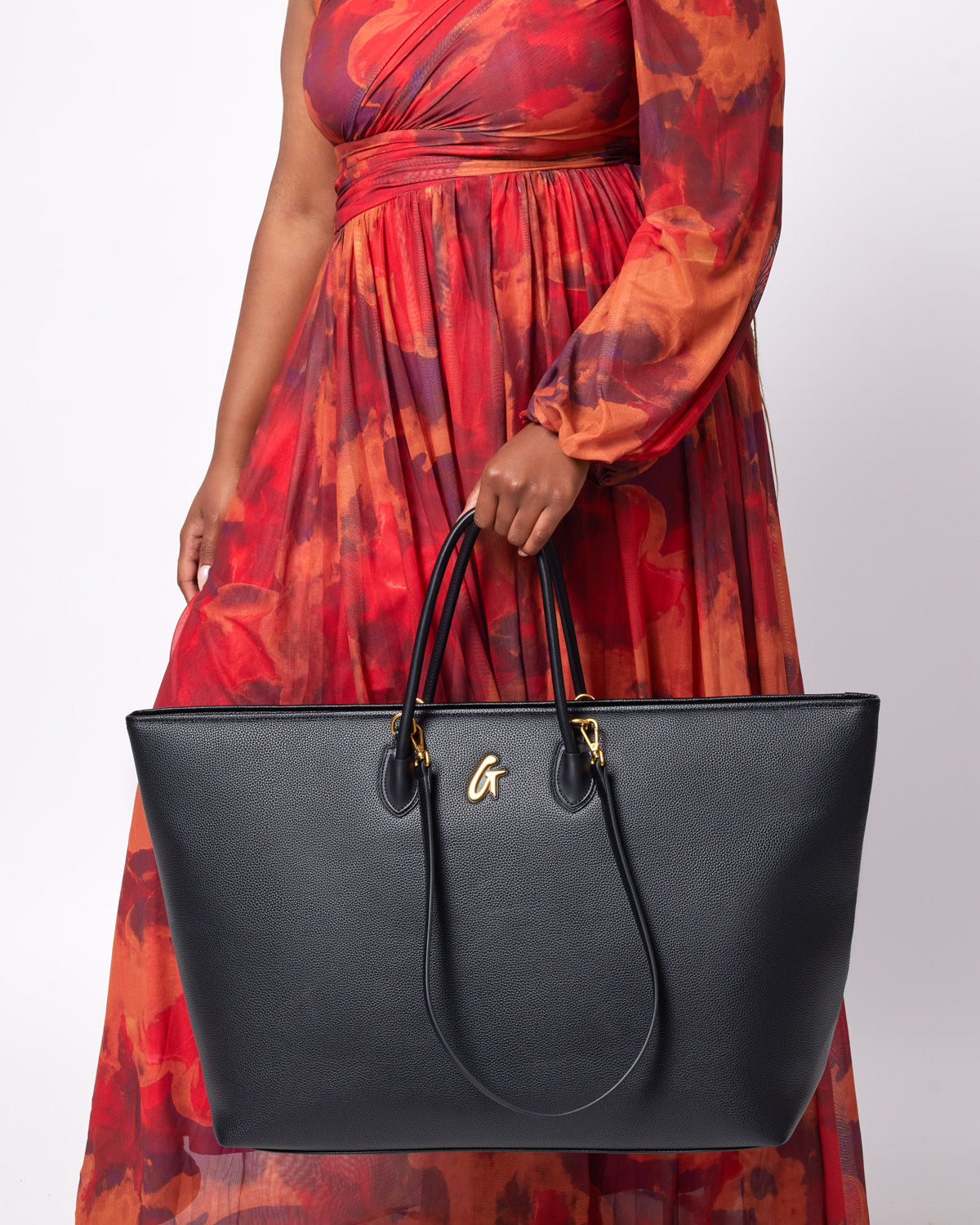 Black jumbo pebble tote bag with gold accents, held by a woman in a colorful red-orange dress.